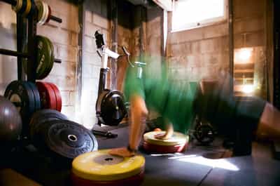 Dynamic motion blur capture of person exercising in home garage gym with colorful Rogue weight plates, natural lighting streaming through wooden structure, conveying movement and fitness energy on Aerocolor IV film