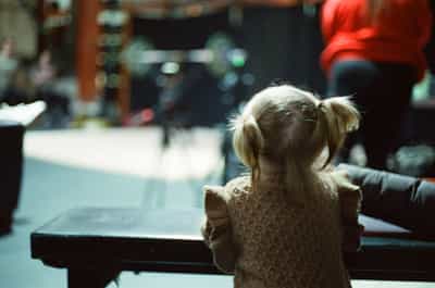 Tender back-of-head portrait of toddler with blonde pigtails in beige sweater watching Olympic weightlifting competition from barrier railing, with blurred gym environment and colorful bokeh lights from competition setting creating dreamy background, captured on CineStill 800T film with cinematic tungsten color palette and distinctive halation effects, evoking the next generation of athletes and the family atmosphere of competitive weightlifting culture