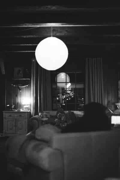 Atmospheric black and white interior photograph of living room at night featuring glowing spherical paper lantern pendant light suspended from exposed beam ceiling, illuminating vintage wood furniture and decorative objects, with blurred Christmas tree lights visible through multi-pane window creating bokeh in background, dog resting in soft-focused foreground, captured on Ilford HP5+ 400 film with dramatic contrast between bright lantern and deep shadows, evoking cozy winter evening solitude and contemplative domestic tranquility with timeless monochrome aesthetic
