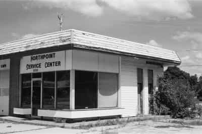 Classic mid-century Northpoint Service Center building with weathered white siding, large plate glass windows, and distinctive angular roofline under dramatic cloudy sky, capturing small-town Americana on Ilford HP5+ 400 black and white film