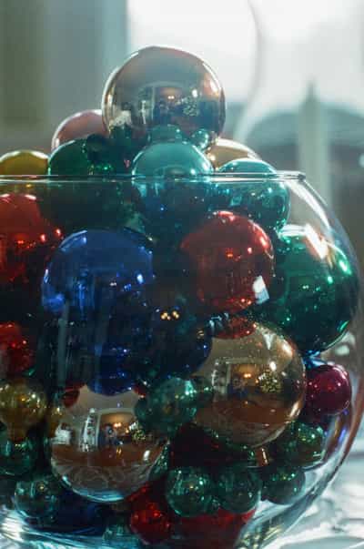 Creative macro composition of colorful vintage Christmas ornaments including emerald green, sapphire blue, ruby red, and gold mercury glass baubles piled in clear glass bowl, with photographer's distorted reflection visible in spherical ornament surfaces creating meta self-portrait, soft window light illuminating translucent glass with rich jewel tones and highlights, captured with shallow depth of field on Kodak Ektachrome E100 slide film with saturated vintage color palette and fine grain, evoking nostalgic holiday traditions and contemplative artistic documentation