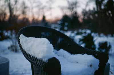Moody atmospheric scene of dark wicker patio chair covered with thick layer of pristine white snow in foreground with blurred snow-dusted furniture and winter garden beyond, captured during blue hour twilight with soft cool tones and shallow depth of field creating dreamy bokeh effect, shot on Kodak Ektachrome E100 slide film with muted winter color palette featuring deep blue shadows and crisp white highlights, evoking quiet solitude of winter evenings and the peaceful beauty of snow-blanketed outdoor spaces awaiting spring
