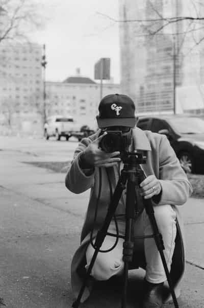 Dynamic street-level portrait of a photographer crouching on an urban sidewalk while composing a shot through a camera mounted on a sturdy tripod, wearing a dark baseball cap with embroidered logo and light-colored zip hoodie, face obscured behind the camera body in a moment of focused creative intent, bare winter trees and mid-rise city buildings with parked vehicles lining the street visible in the soft-focused background, shot on Ilford HP5+ 400 black and white film with rich tonal contrast between the deep blacks of the cap and camera and the bright highlights of the overcast sky, moderate grain adding gritty urban texture to this candid documentary-style capture of a fellow photographer at work