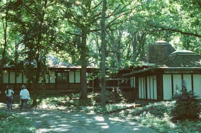 Two people walking toward modernist ranch-style house with horizontal glass windows and natural wood siding nestled among dense mature forest canopy, creating harmony between contemporary architecture and wooded landscape, captured on CineStill 800T film with warm natural lighting