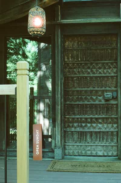 Detailed portrait of a dramatically carved geometric wood front door framed by the mid-century modern home's signature dark trim, with an intricate Moroccan-style pierced metal pendant lantern glowing warm amber overhead and casting a small red halation bloom characteristic of tungsten-balanced film stock, rough wooden porch post in the foreground, lacquered 'OPEN SUNDAY' sandwich board sign leaning against the post, welcome mat at the door's base, lush green forest reflected in the adjacent floor-to-ceiling window, cool teal and green tonal palette throughout, captured on CineStill 800T film with its distinctive cinematic color rendering evoking architectural reverence and the ritual of arrival at a singular home