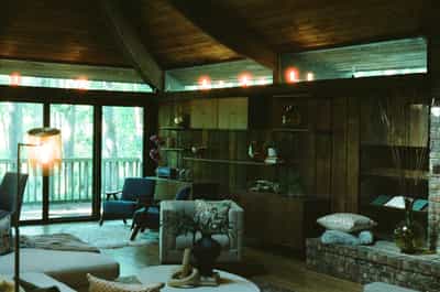 Stunning mid-century modern living room interior featuring exposed wooden beam ceiling, floor-to-ceiling windows with forest views, brick fireplace, contemporary furniture, and warm ambient lighting with distinctive CineStill 800T halation effects around lamps creating cinematic tungsten atmosphere