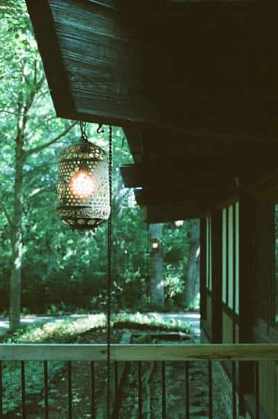 Atmospheric covered porch scene featuring ornate perforated metal lantern hanging from wooden ceiling with warm tungsten glow, surrounded by lush forest foliage visible through columns, captured on CineStill 800T film with cinematic lighting and distinctive halation effects creating moody woodland retreat ambiance
