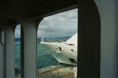 Architectural framing of a cruise ship's sleek white prow viewed through a weathered pale concrete dockside archway, massive cylindrical support columns bracketing the composition on either side, taut white mooring lines descending in parallel diagonals from ship to unseen bollards below, the vessel's dark-windowed navigation bridge and curving hull geometry emphasized against a dramatic broken sky of cumulus shadow and small patches of sunlit Caribbean sea beyond, a small dockworker figure in fluorescent orange barely visible at the distant pier level providing critical scale, pale distant headland forming a soft silhouette on the horizon, captured on Kodak Ektachrome E100 slide film with moody layered composition evoking the heavy architectural presence of modern ocean liners at dock