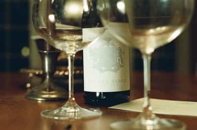 Elegant wine still life featuring Signal Ridge Pinot Noir bottle with embossed heraldic crest label positioned between two crystal wine glasses filled with golden wine on rustic wooden table, captured with shallow depth of field and warm ambient restaurant lighting, shot on CineStill 800T film with cinematic tungsten color palette and distinctive halation glow creating intimate evening dining atmosphere with rich bokeh and vintage aesthetic