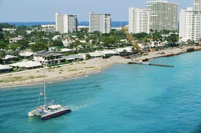 Sweeping elevated perspective from cruise ship altitude of a brightly painted catamaran motoring across impossibly turquoise inlet waters off Fort Lauderdale's beach spit, with a thin band of low-slung beachfront houses, mature palms, and a lone yellow construction crane forming the middle ground before a clean horizon line of pale high-rise condo towers stretching the length of the coast, captured on Kodak Ektachrome E100 slide film with the stock's signature crisp cyan-and-aquamarine water rendering, deep saturated blue sky, and clean white architectural highlights, the boat anchoring the composition's lower-left third against generous negative space of water and sky.