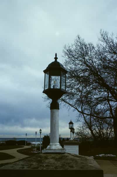 Moody twilight scene of ornate colonial-style street lamps with white pillars and black lantern housings arranged in perspective along waterfront park pathway, bare winter trees silhouetted against overcast blue-gray sky with distant water visible on horizon, captured during blue hour on Kodak Ektachrome E100 slide film with muted cool tones and soft natural light, evoking melancholic New England coastal town atmosphere and the quiet solitude of late winter afternoons by the water