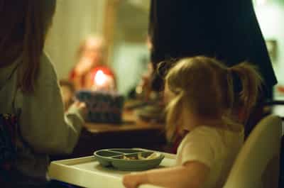 Tender intimate scene of young blonde toddler seated at white children's tray with sectioned plate during family meal, with blurred adults visible in warm softly-lit background featuring glowing candlelight creating bokeh, capturing authentic moment of childhood during holiday gathering with natural documentary style and shallow depth of field, shot on Kodak Ektachrome E100 slide film with warm amber tungsten color palette and soft film grain, evoking nostalgic family traditions and the quiet wonder of early childhood during festive celebrations