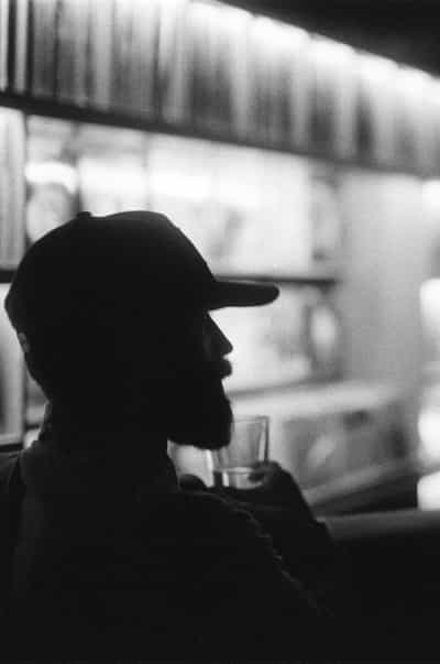 Intimate black-and-white profile silhouette of a bearded man in a backwards baseball cap rendered almost wholly in shadow against a softly glowing wall of vertically stacked vinyl records receding into bokeh behind him, the curved tip of his beard and the line of his nose just catching a faint edge of warm light as he holds a low rocks glass of clear liquid up near his chest, the geometric brackets of the illuminated record shelving glowing in horizontal bands above his cap, captured on Ilford HP5+ pushed two stops in development for the deep velvet blacks, lifted highlights, and gritty silver-grain texture that defines the nocturnal atmosphere of the Wiggle Room record bar in downtown Milwaukee