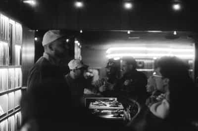 Documentary black-and-white scene of a tight gathering inside the Wiggle Room record bar in downtown Milwaukee, a man in a white backwards cap and dark shirt standing in profile at the left edge of the frame anchoring the composition as he turns toward a clustered group of friends in caps and dark clothes gathered around the DJ booth at center-right, drinks raised mid-conversation, a turntable and mixer in cardboard packaging set on the bar between them, the long horizontal glow of a backlit liquor shelf and neon ceiling strip washing the rear wall, the towering wall of softly illuminated vertical vinyl spines glowing along the left edge, captured on Ilford HP5+ pushed two stops in development for rich blacks, blown speculars, and the unmistakable grain structure of available-light film documentation