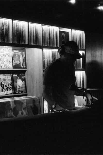 Classic black-and-white DJ portrait of a young man in a baseball cap and over-ear headphones standing in concentration behind a pair of turntables at the Wiggle Room record bar in downtown Milwaukee, his silhouetted torso and the curve of his shoulders backlit by the radiant grid of illuminated vinyl spines glowing in horizontal racks behind him, a few featured record sleeves propped face-out below the main wall, one hand resting on the deck and the other near a control as he cues the next track, captured on Ilford HP5+ pushed two stops in development for the punchy contrast, lifted shadows, and silver-rich grain that suits a working portrait of a DJ in his element
