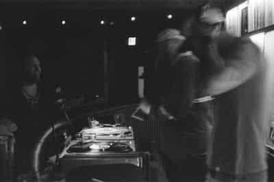 Loose available-light black-and-white documentary frame of the DJ booth in motion at the Wiggle Room record bar in downtown Milwaukee, a laughing man on the far left standing crisp behind a microphone while two figures on the right blur dramatically across the frame mid-movement — one in a white cap holding a record sleeve, another with arms raised — the turntable platter and a vinyl record spinning sharply in the center-left, a softly glowing emergency exit sign and overhead spotlights stretching across the dark ceiling, captured on Ilford HP5+ pushed two stops in development for the high contrast, kinetic motion smear, and grainy nocturnal energy of a live DJ set on film