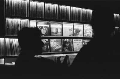 Bold black-and-white environmental study of the Wiggle Room record bar's signature wall of glowing backlit vinyl racks in downtown Milwaukee, two silhouetted heads and shoulders rising along the bottom edge of the frame as patrons lean toward the displayed record sleeves, the upper shelf rows packed with tightly stacked vinyl spines illuminated from behind, the middle rows featuring face-out album covers including Idris Muhammad's Boogie to the Top, Wu-Tang Clan's C.R.E.A.M., a Dr. Dre sleeve, and the Sex Pistols' Never Mind the Bollocks, the lower bins of flipping crates barely visible in the shadowed foreground, captured on Ilford HP5+ pushed two stops in development for deep velvet shadows, glowing highlight halos, and the rich silver grain that turns a record-store wall into iconic black-and-white documentary