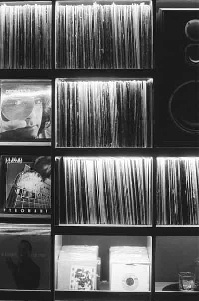 Tightly composed black-and-white study of the Wiggle Room record bar's illuminated vinyl shelving in downtown Milwaukee, three stacked rows of vertically slotted record spines glowing softly from internal lighting and filling most of the frame in a precise geometric grid, a large round studio monitor speaker mounted along the right edge, two featured face-out album sleeves on the left including Def Leppard's Pyromania anchoring the middle row, a bottom shelf of small singles and 45 sleeves with a clear rocks glass tucked into the lower-right corner, the faint silhouette of a patron just visible in reflection on a lower sleeve, captured on Ilford HP5+ pushed two stops in development for crisp tonal separation between the bright record edges and the surrounding shadow architecture of the bar