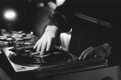Intimate black-and-white close-up of a DJ's hands working a vinyl record on a turntable at the Wiggle Room record bar in downtown Milwaukee, one hand resting flat across the spinning platter to cue the record while the other hand rests on the mixer with a glint of a wedding band catching the warm overhead lamp light, the dark sleeves of a long-sleeve shirt anchoring the right two-thirds of the frame, the soft glow of a single tungsten lamp blooming in the upper left and a second deck with a crate of records receding into shadowed bokeh behind, captured on Ilford HP5+ pushed two stops in development for velvet blacks, blooming highlight on the chrome platter edge, and the silver-grain texture that defines tactile film documentation of analog music being played