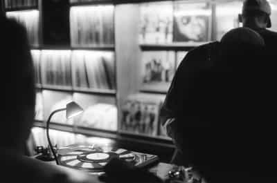 Atmospheric over-the-shoulder black-and-white frame from inside the DJ booth at the Wiggle Room record bar in downtown Milwaukee, a goose-necked tungsten desk lamp on the left side of the frame casting warm directional light onto the turntable's spinning platter as a hand reaches toward the deck in the lower foreground, the broad silhouetted back of the DJ in a dark shirt occupying the right side, a baseball-capped patron leaning in just behind, the glowing wall of stacked vinyl spines and featured record sleeves filling the background beyond the booth in soft focus, captured on Ilford HP5+ pushed two stops in development for the deep shadowed booth interior, lifted record-wall highlights, and gritty silver grain of a working-night document on film