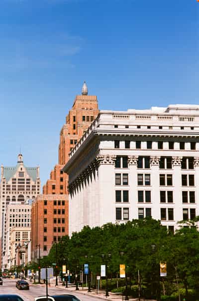 Downtown Milwaukee streetscape looking along Wisconsin Avenue, a monumental white classical-revival building with a heavily corniced, column-articulated facade and rows of dark punched windows filling the right of the frame while a cluster of older red-brick towers — one capped by a stepped, ornamented crown — recedes into the mid-distance, leafy green street trees, lamp posts and colorful banners lining the avenue below and a single car on the sunlit roadway, all beneath a clear gradient blue sky, captured on Kodak Ektar 100 whose vivid saturation and fine grain sharpen the contrast between pale stone and warm brick and give the urban canyon a clean, postcard-bright vibrancy.